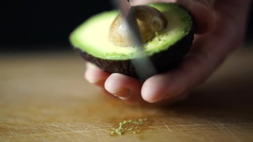 Caucasian male hand holding halved avocado and taking out its pit with sharp chefs knife, SLOW MOTIO