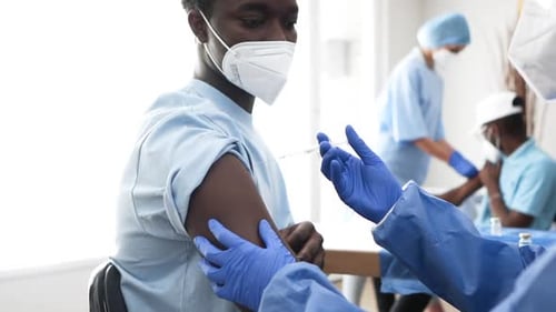 Nurse Administers Covid-19 Vaccine Shot In Left Arm Of Black Man Wearing Medical Mask In The Clinic.