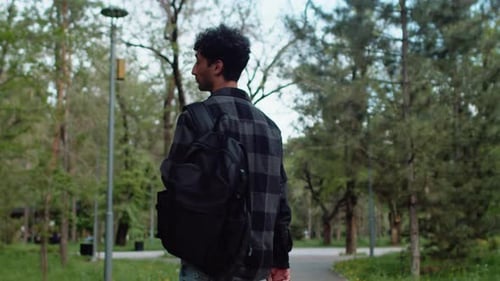 Young Curly Man in Casual Clothes with a Backpack Walking in an Alley of the City Park Then Turning