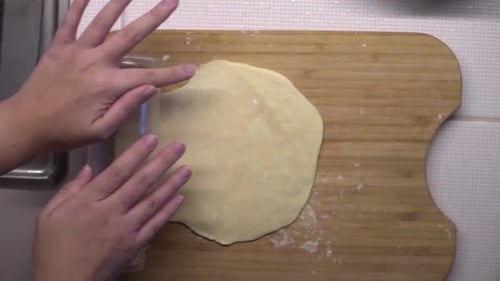 Hands rolling thin dough with plastic bottle on wooden cutting board, overhead shot