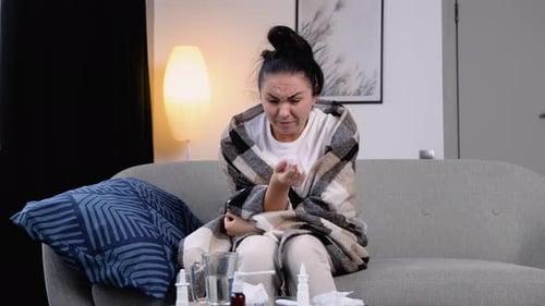 Young Woman Sits on the Couch at Home Under a Plaid Sneezing in a Napkin Feels Weak Unwell Needs