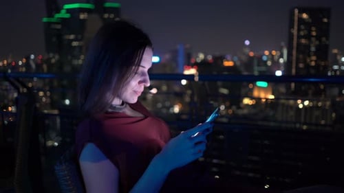 Happy young woman texting on smartphone at rooftop bar in city nightlife