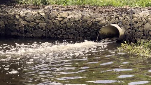 Water rapidly flows from a large pipe into a river or stream. The water is murky and turbulent as it