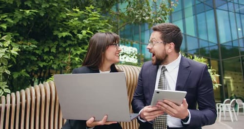 Joyful businessman and businesswoman in formal attire sitting on bench outside modern glass office