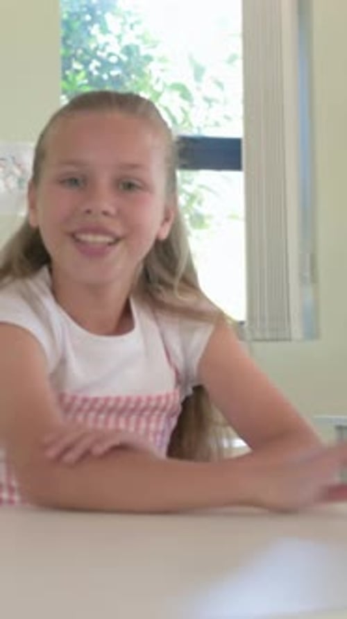 Vertical video: Reacting to cue girl smiling, reaching at desk in classroom, wearing pink pinafore