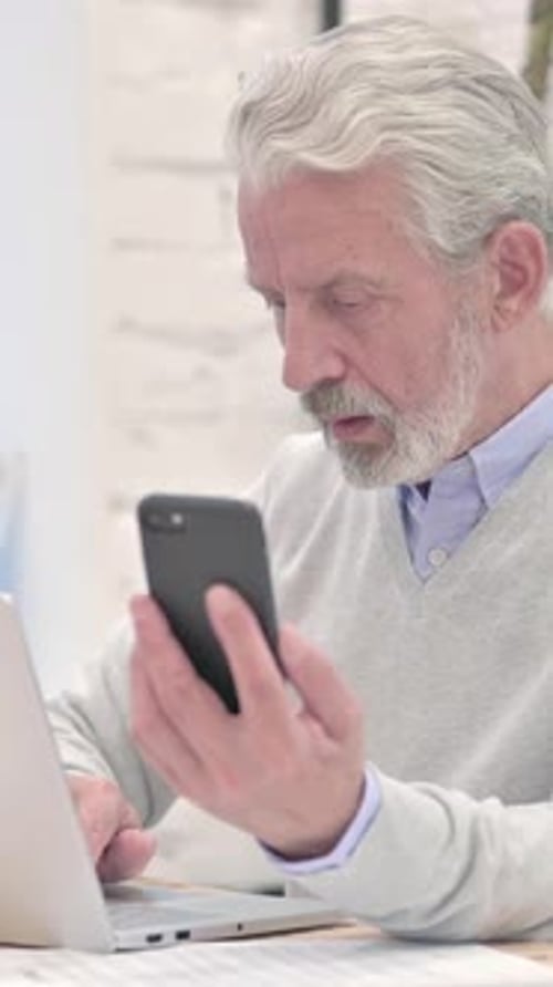 Senior Man Using Smartphone with Laptop in Office