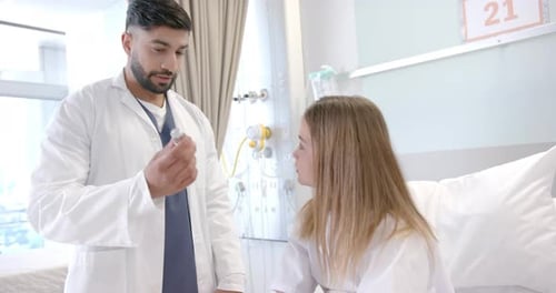 Diverse male doctor showing girl patient in hospital bed how to use asthma inhaler, slow motion