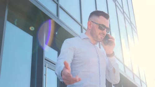 A Man in Dark Glasses is Gesturing While Talking on a Mobile Phone in Front of an Office Building