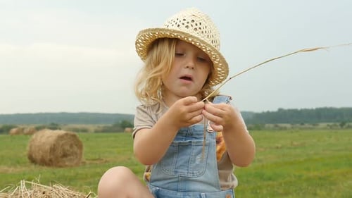 Funny Child Sitting On Haystack On Farmer Field. Adorable Little Girl Is Smiling