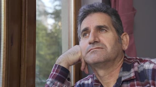 The man is at home watching out the window and is thoughtful.