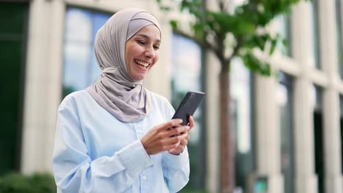 Close up. Young smiling muslim female employee in hijab is using a smartphone standing on the street