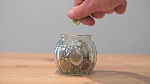 A close-up of a hand dropping a coin into a glass jar filled with coins, symbolizing saving money, f