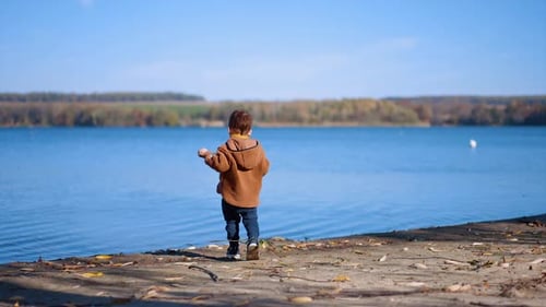 Baby boy spending sunny autumn day at the river. Caucasian toddler throwing the stones into water.