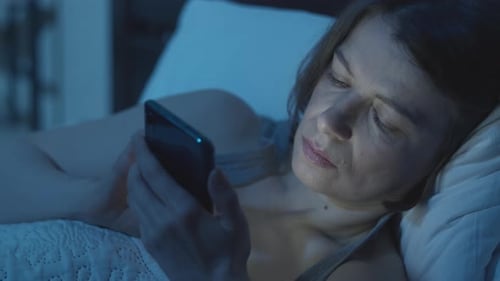 A woman uses a mobile phone while lying in bed in a dark room.