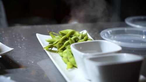 Steaming pile of edamame in restaurant prep kitchen plated on white dish, slow motion orbit HD