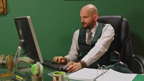 A manager sits behinds his computer and scrolls with the mouse. He types down a few notes and grabs