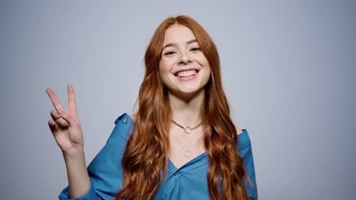 Mulher ruiva alegre faz gesto de mão de sinal de paz no fundo cinza do estúdio