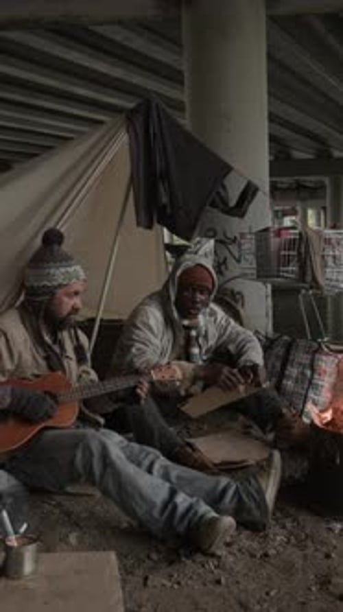 Two Beggarmen Sitting on Ground under Bridge by Fire, One Playing Guitar