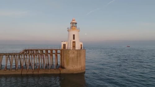 Scenic lighthouse, calm sea and jetty at golden hour, with a full moon and boat in background