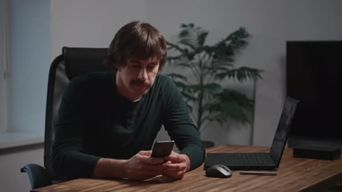 Man Using Mobile Phone at Desk with Laptop