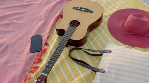 A guitar lies next to a hat and phone on a beach blanket