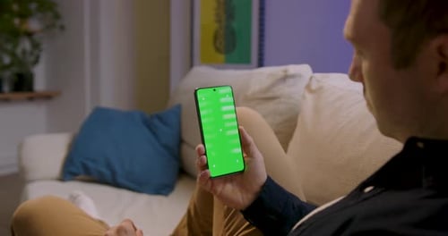 Man Holding Cell Phone With Green Screen While Resting On The Couch At Home. - close up shot