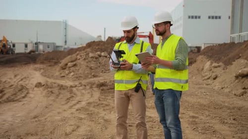 Two Builders in Bright Yellow Vests Holding Tablet and Smartphone While Controlling Flying Drone