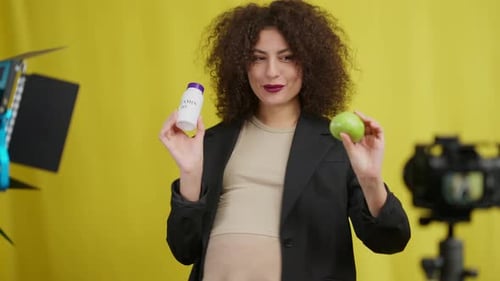 Smiling Pregnant Woman Showing Green Apple and Bottle of Vitamin Supplements Filming Vlog at Yellow