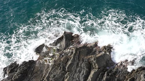 Top down aerial view of waves crashing on rocky shoreline. Blue ocean water on steep cliffs.