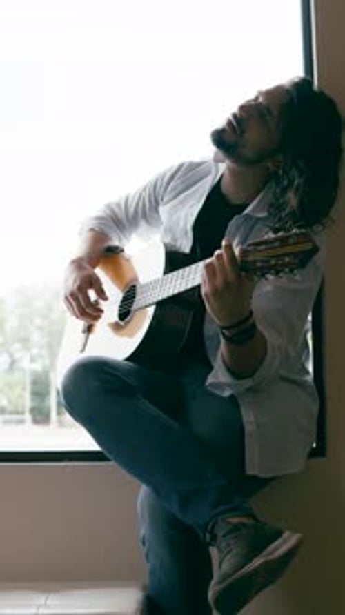 Man Plays Guitar by Window, Relaxing at Home