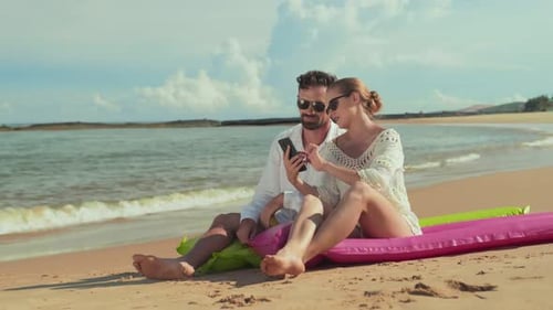 Young Couple Talking and Looking Through Photos in Smartphone on the Beach