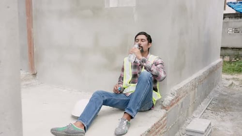 Man wearing green reflective vest taking off helmet Sitting against a cement wall after working on a