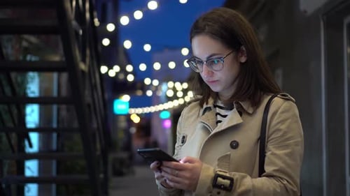 A Young Woman Chats on a Smartphone Late at Night on a Narrow Street A Girl in Glasses with a Phone