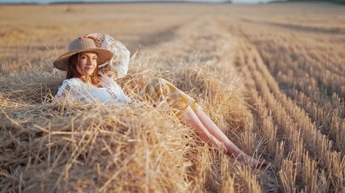 Pretty Woman in Straw Hat and Embroidered Blouse Lying on Hay in Countryside at Sunset