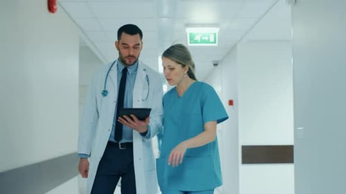 Surgeon and Nurse Walk Through Hospital Hallway, They Consult Digital Tablet Computer while Talking