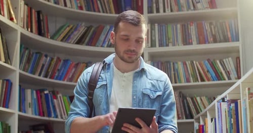 Caucasian Young Male Student Standing in Library Using Tablet Device and Tapping on Screen Stylish