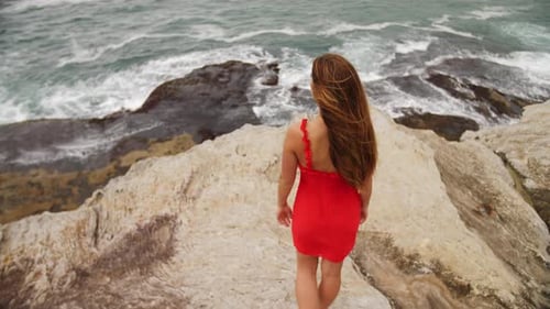 Sexy Girl In Red Dress Standing On The Rock And Looking Up In The Sky With Ocean Waves In The Backgr