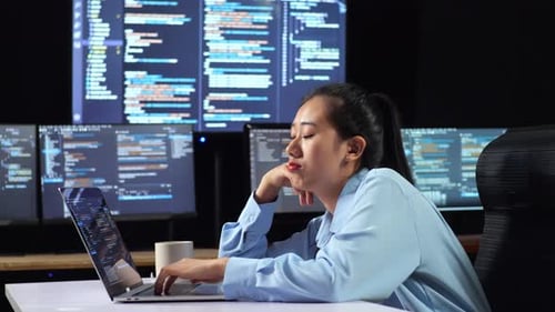 Vista lateral de una programadora asiática aburrida escribiendo código con una computadora portátil con varios monitores