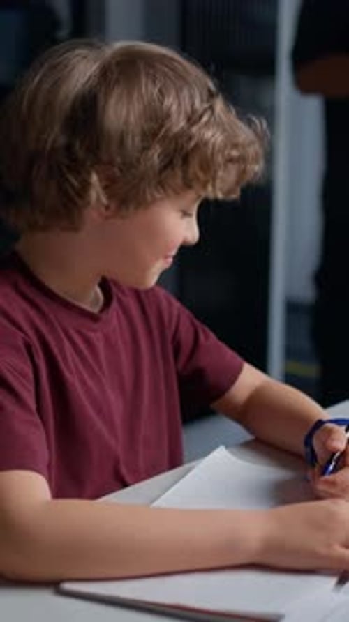 Young Boy Focused on Writing in a Notebook with Pen While Seated at a Desk Indoors Accompanied By an
