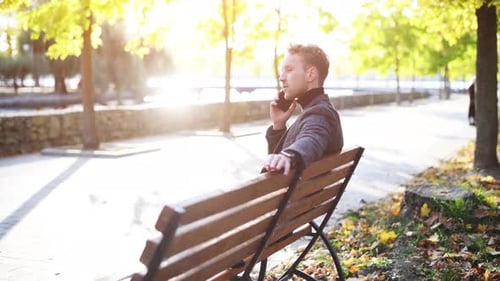 Young man in formal clothes sitting in the autumn park at sunny day have a call