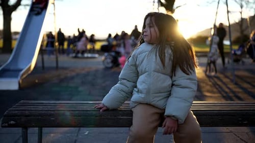One contemplative little girl sitting at park bench wearing coat during winter sunset time with