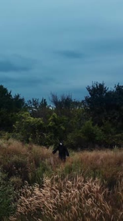 Mysterious ghost in black cloak among nature. Dark figure standing on the field under cloudy sky.