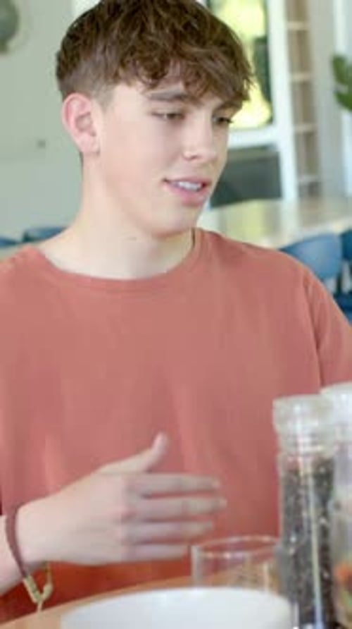 Vertical video: Noticing cue, teen in orange tee turning, gesturing at kitchen table with glass