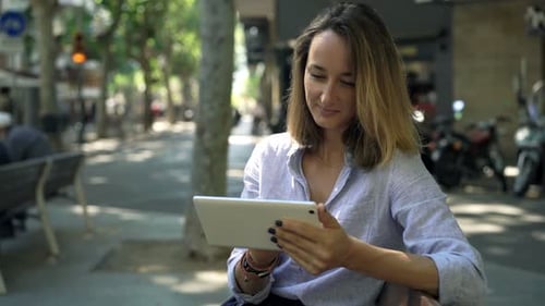Happy young businesswoman with tablet sits on bench in the city smiling