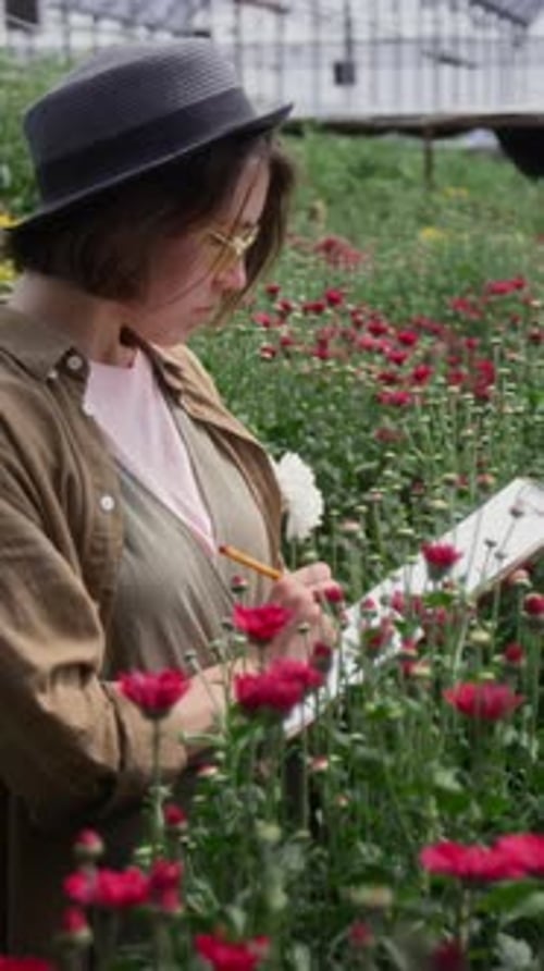 Woman Inspecting Flowers and Taking Notes in Garden