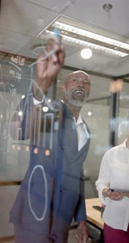 Vertical video of african american businessman writing on glass wall in office with copy space
