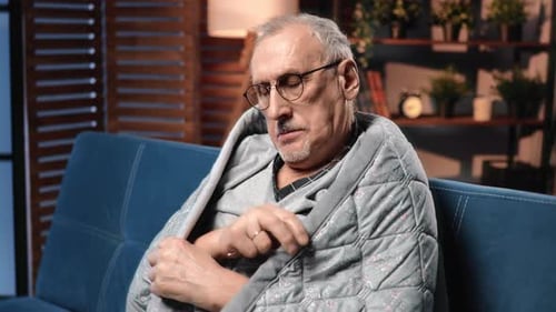 Senior Man Sitting on Couch Wrapped in Quilt