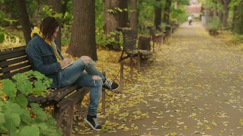 Young Creative Man with Dreadlocks Drawing Writing in Notebook Sitting on Park Bench in Golden