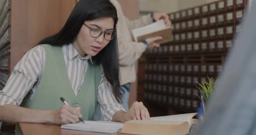 Intelligent Asian Girl Studying in Library Preparing for University Exam Writing and Reading Book