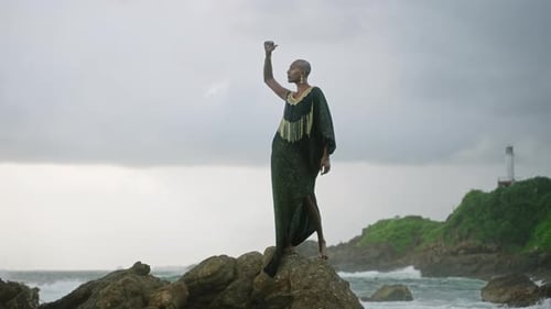 Androgynous Person of Color Stands in Boutique Posh Dress on Rock Above Ocean Coastline Trans Sexual